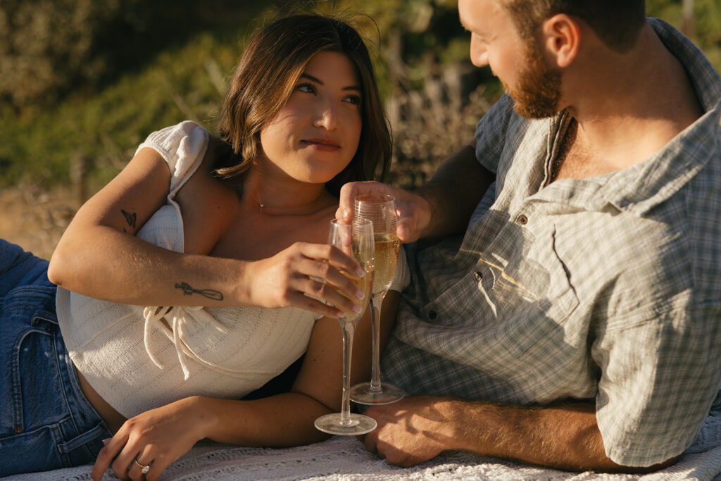 A couple in Southern California cheers with  glasses of champagne on a picnic blanket during their engagement pictures san diego