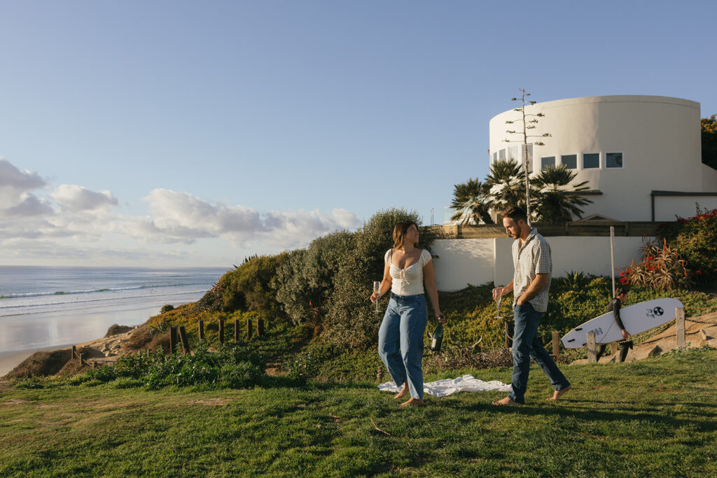 A surfer carries his board in the background of a pacific beach photography session for an engaged couple 