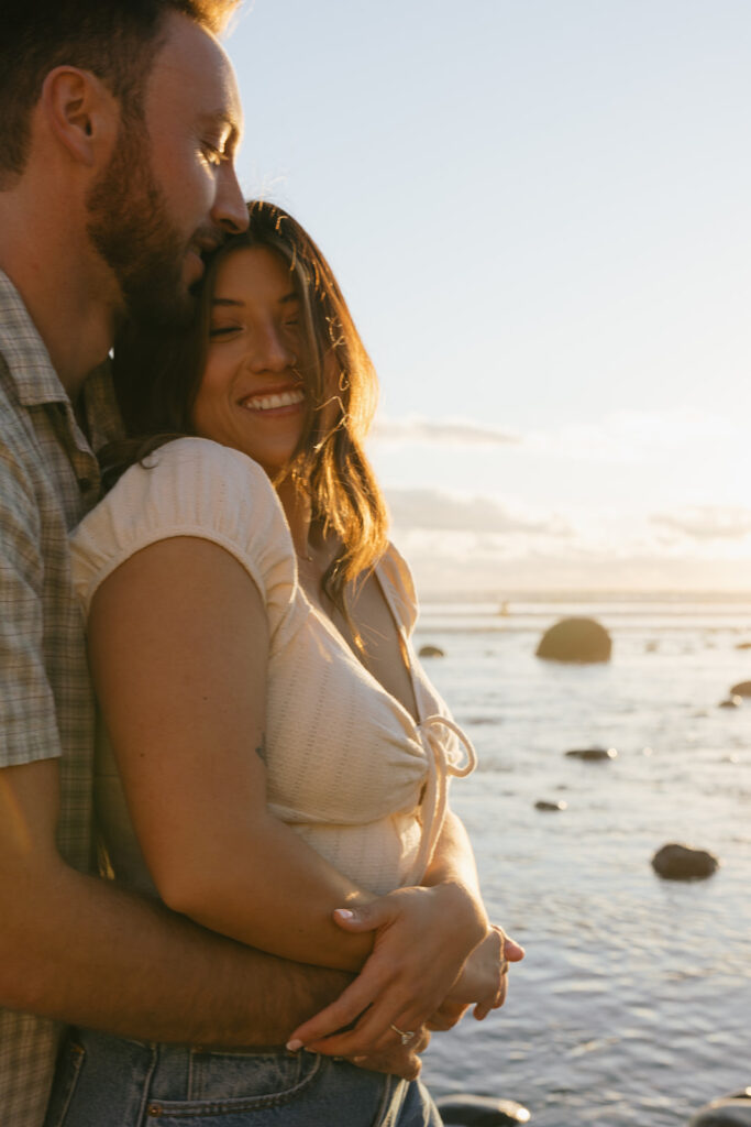 A man wraps hugs his fiancée from behind with views of a Pacific sunset in the background during their San Diego engagement photos 