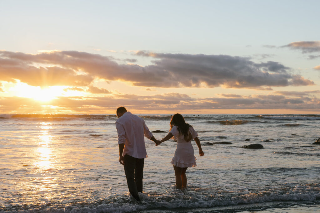 A California couple holds hands as they wade into the Pacific ocean during their San Diego engagement photoshoot 