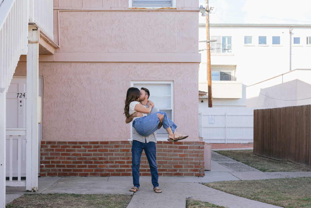 A man wearing jeans & flip flops holds his fiancée in his arms as they share a kiss outside a stucco apartment building during their san diego engagement photos with Allison Nicole Photo 