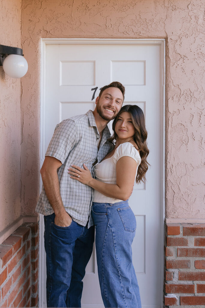 A CA couple smile and embrace outside an apartment door in Southern California during their engagement photos san diego