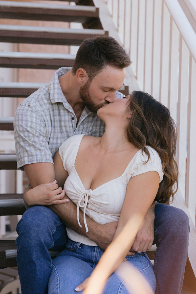 A woman in a white cap sleeve top and jeans kisses her fiancé wearing a plaid shirt on the steps of his former apartment building during their engagement photos in san diego 
