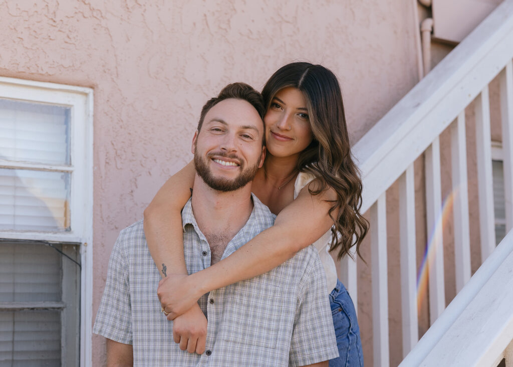A woman with wavy brown hair wraps her arms around her fiancés neck outside a San Diego apartment building during their california engagement photos 