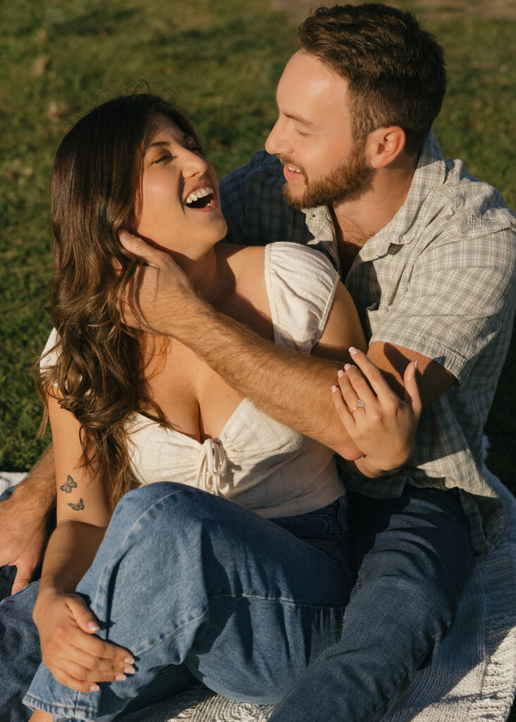 A newly engaged couple smile at each other atop a picnic blanket near Tourmaline Beach during their San Diego engagement photos 