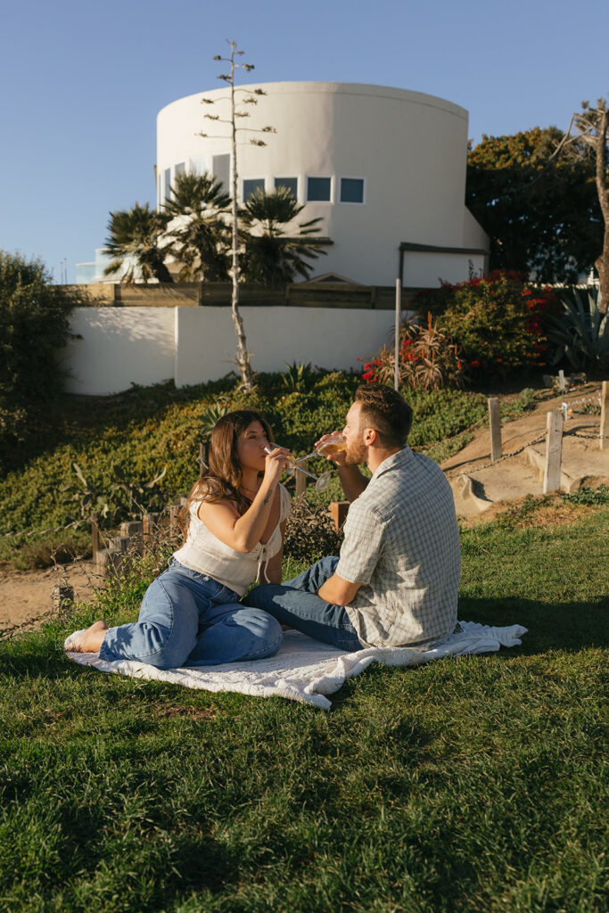 A Southern California couple share celebratory glasses of champagne on a picnic blanket during their san diego engagement photography session